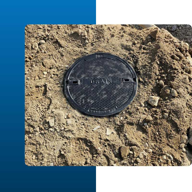 black cast iron manhole cover embedded in a mound of light brown dirt and gravel. The word "DRAIN" is clearly embossed in the center of the textured surface, along with the "Made in USA" stamp at the bottom.