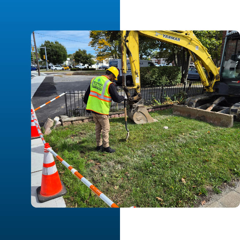 A construction worker wearing a yellow hard hat and a high-visibility safety vest stands on a grassy lawn next to a yellow Yanmar mini-excavator. The worker is holding a shovel, preparing to dig or mark a spot near the excavator's bucket.