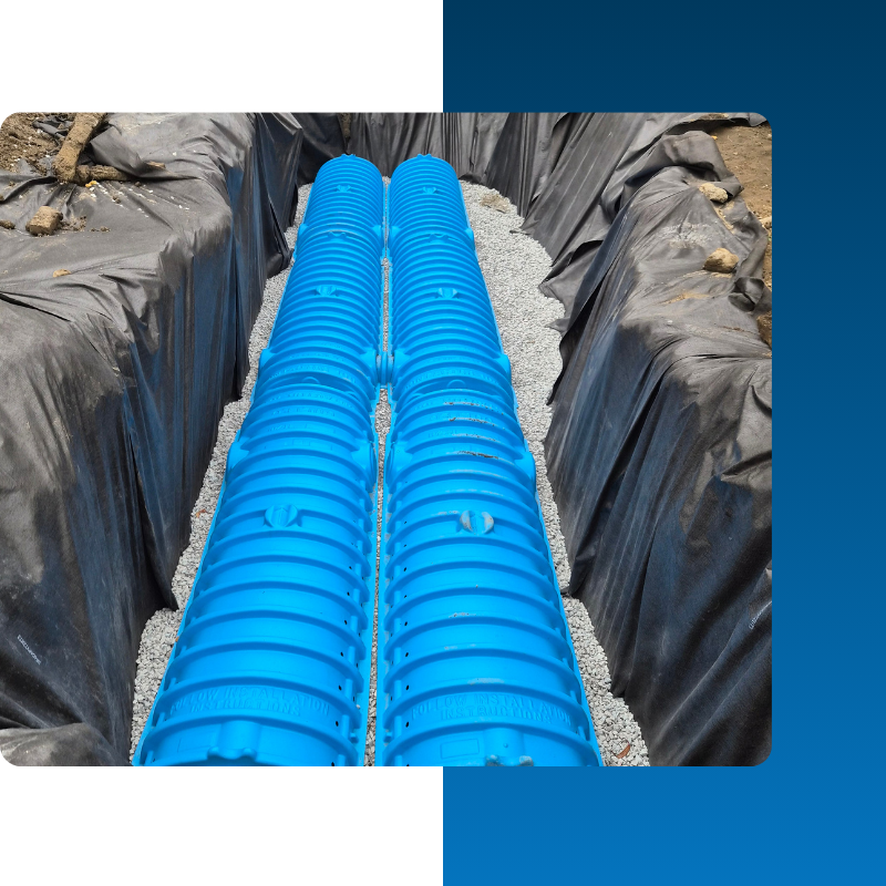 An underground installation of two parallel rows of bright blue, arched plastic drainage chambers (leach field chambers) set in a trench. The chambers are surrounded by a layer of grey crushed stone and the trench is lined with a black geotextile fabric.