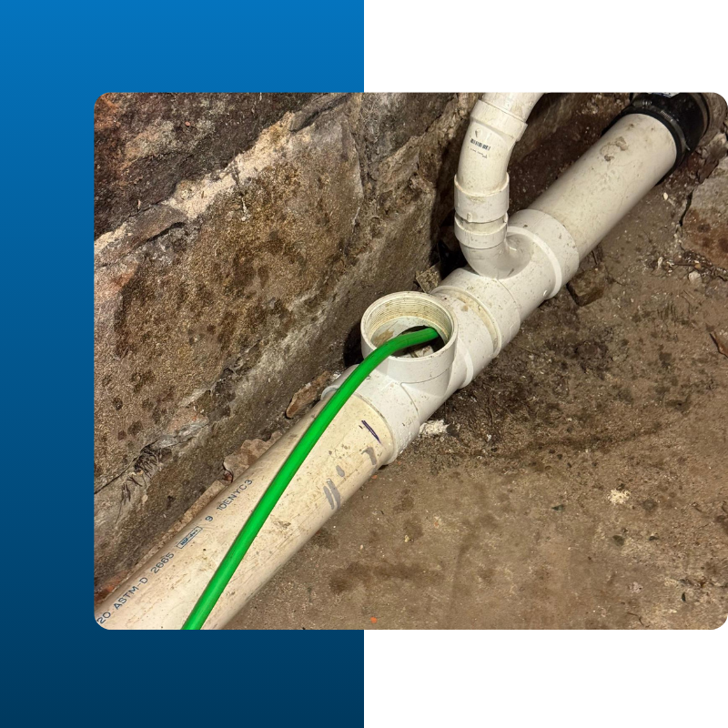 A bright green inspection camera cable is inserted into an open floor drain or toilet flange in a bathroom with dark wood-style tile flooring