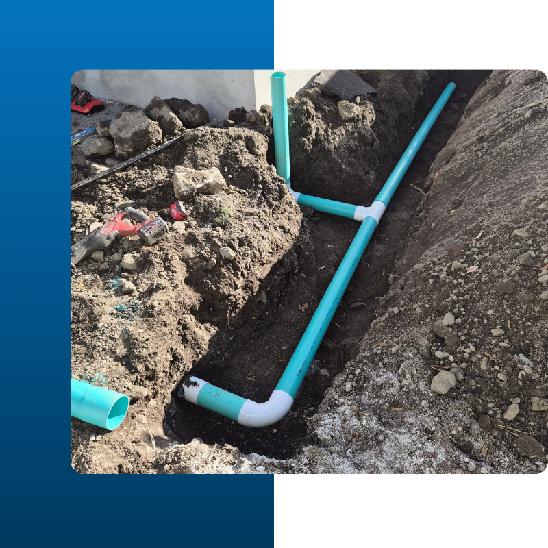 A newly installed teal-colored PVC drainage system sits in a narrow dirt trench next to a concrete building foundation. The piping includes a main line with a T-junction and an elbow joint connected by white fittings.