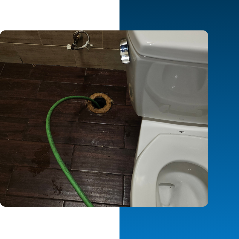 A green sewer camera cable is being fed into an open PVC cleanout pipe located in a basement. The white drain pipe runs along a dirt and concrete floor next to a rustic stone foundation wall. This setup is used for a video pipe inspection
