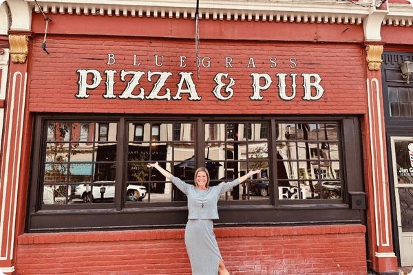 A woman stands with her arms outstretched in front of the "Bluegrass Pizza & Pub" storefront, which is housed in a classic red brick building with large black-framed windows.