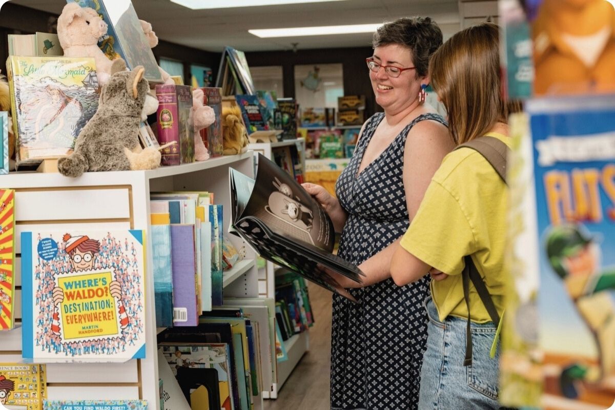 A woman with glasses and a young girl smiling together while looking at an open picture book in a colorful children's section of a bookstore or library.