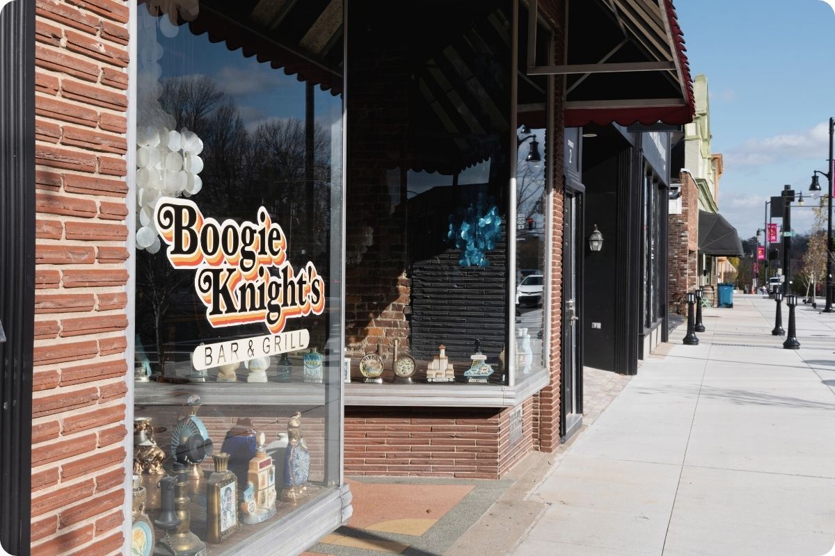 A street-level view of a brick building featuring the "Boogie Knight's Bar & Grill" logo on its front window. The sidewalk extends past several storefronts with black awnings under a bright sky.