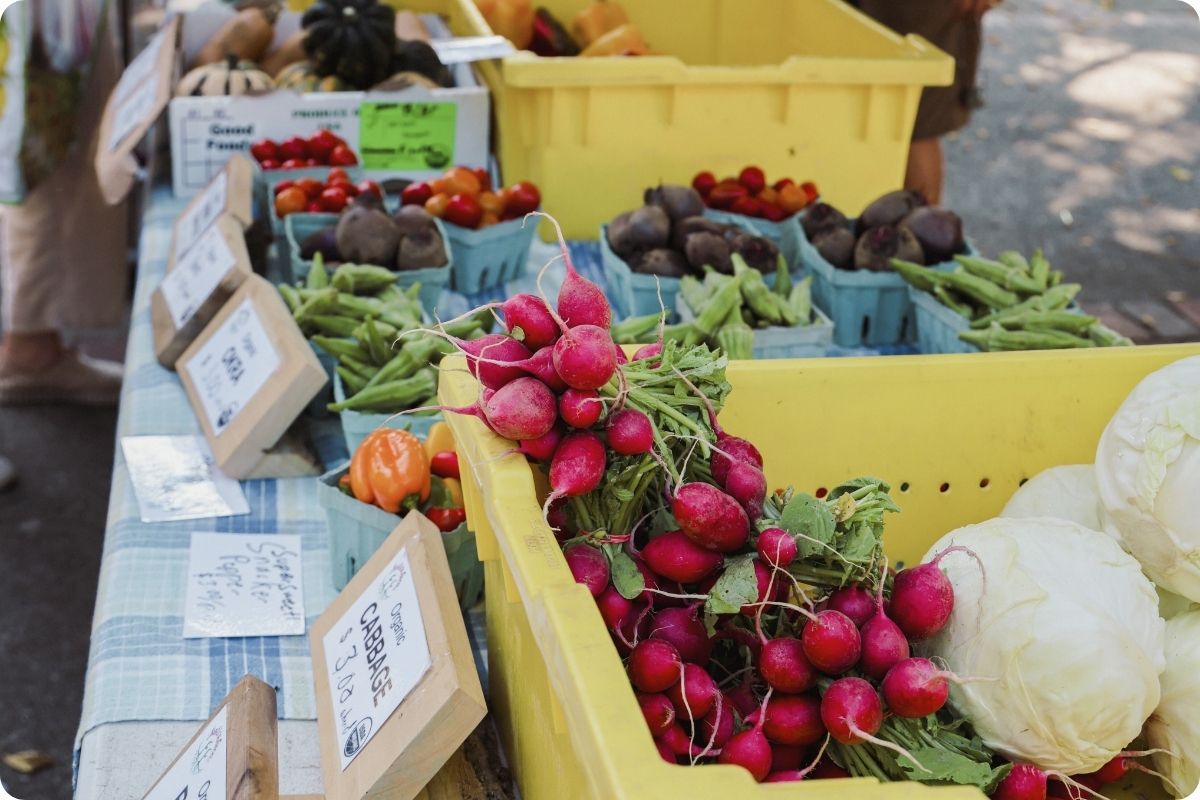 A close-up view of fresh produce at an outdoor farmers market. Bright red radishes with green stems are piled in a yellow bin in the foreground, surrounded by baskets of tomatoes, beets, and okra.