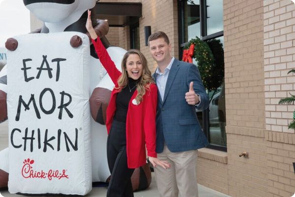 Two cheerful professionals, a man and a woman, standing outside a Chick-fil-A. They are giving a "thumbs up" and waving next to a large inflatable "Eat Mor Chikin" cow.
