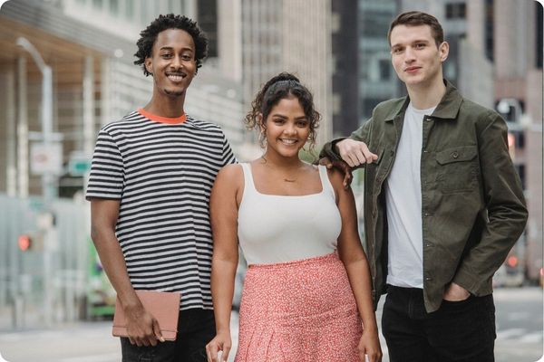 Three young adults stand together on a city street, smiling at the camera. They are dressed in casual, modern clothing with tall city buildings blurred in the background.