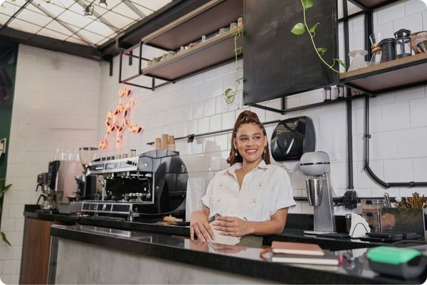 A woman with braided hair smiles warmly while standing behind a modern coffee bar. The background features white subway tiles, a professional espresso machine, and minimalist shelving.