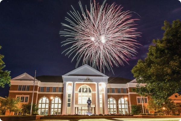 A stunning night shot of Crouse Hall, a large brick building with white columns and arched windows. A vibrant pink and white firework burst illuminates the dark sky directly above the building.