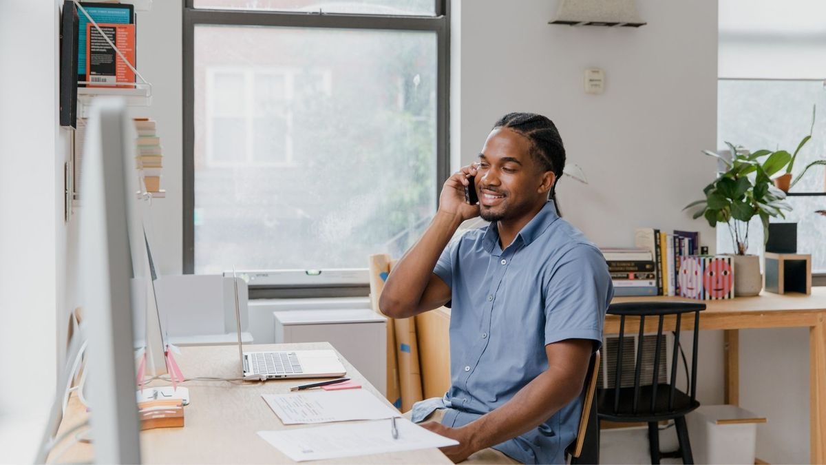 A man in a blue button-down shirt sits at a sunlit desk, smiling while talking on a mobile phone. His workspace includes a laptop, some papers, and a large window in the background.
