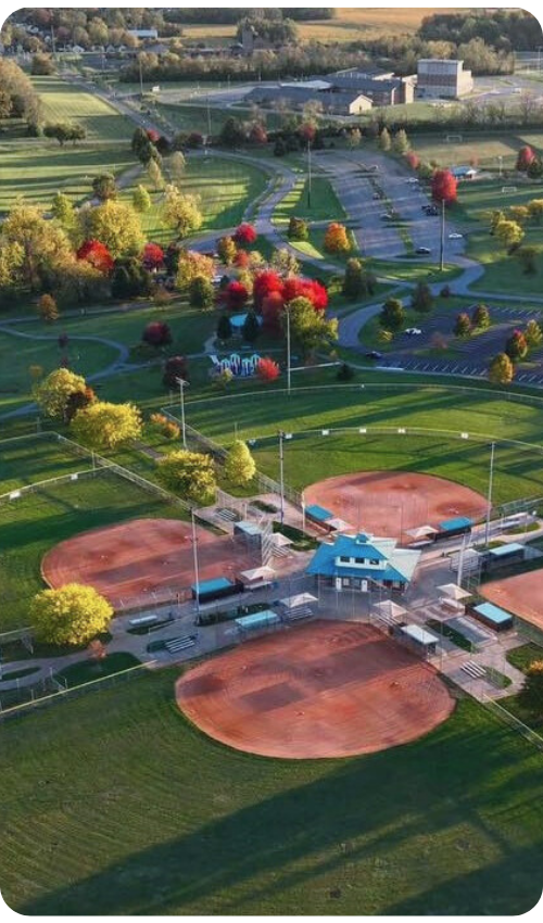An aerial view of a local sports complex featuring several baseball fields surrounded by green space and autumn trees.