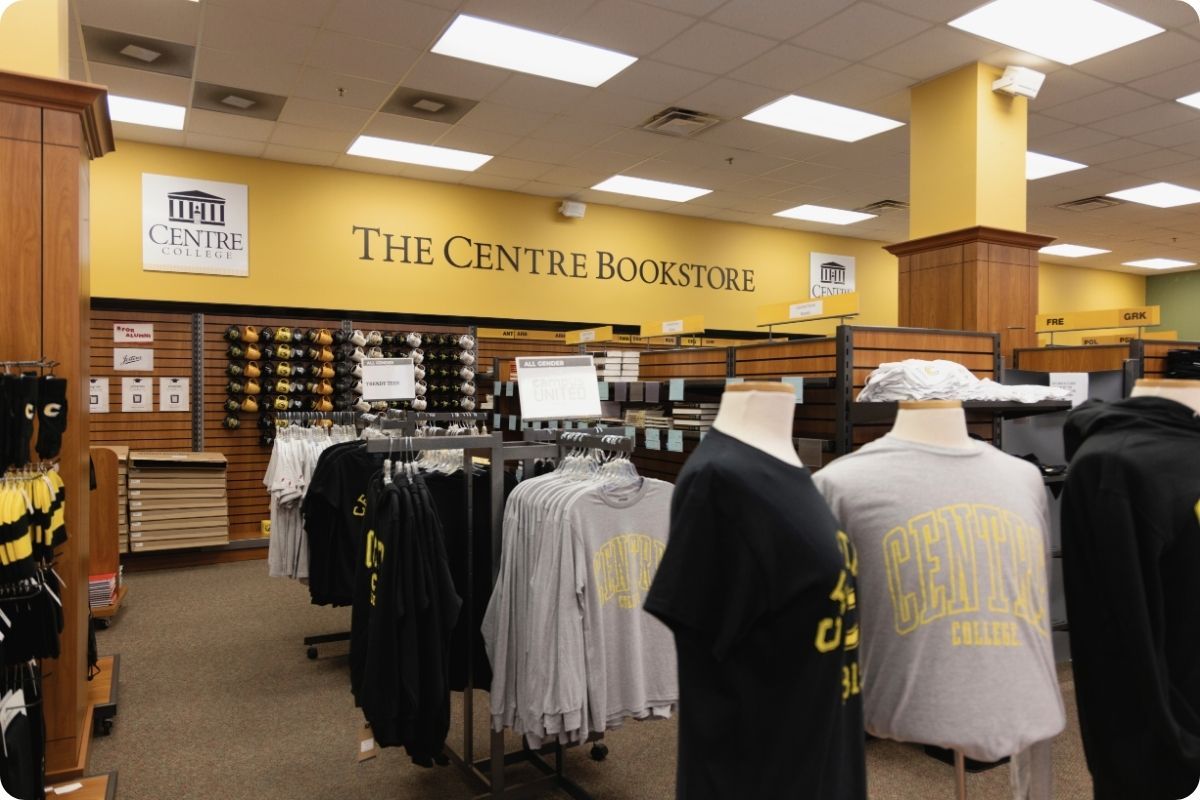 The interior of "The Centre Bookstore," featuring yellow walls and wooden shelving. Clothing racks and mannequins in the foreground display t-shirts and hoodies branded with "Centre College".