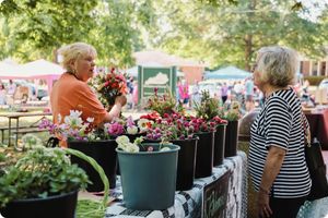 Two women conversing at an outdoor market stall featuring several black buckets filled with fresh-cut, colorful garden flowers.