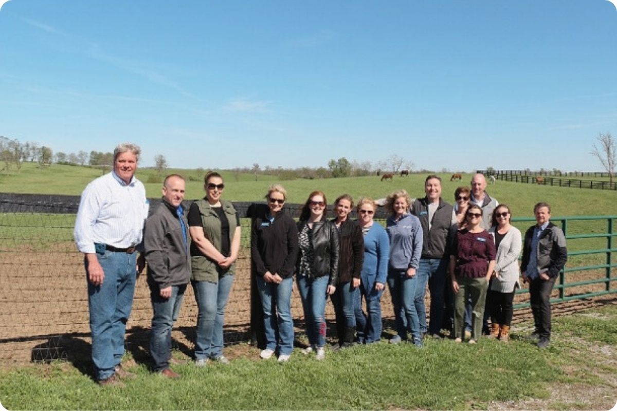 A group of approximately fifteen people in casual business attire stands together for a photo in front of a rustic wooden fence. Behind them is a sprawling green field with several horses grazing under a clear blue sky.