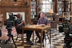 A behind-the-scenes look at a professional video interview. Two men sit at a wooden table in a classic library setting with floor-to-ceiling bookshelves, framed by cameras and production lighting.