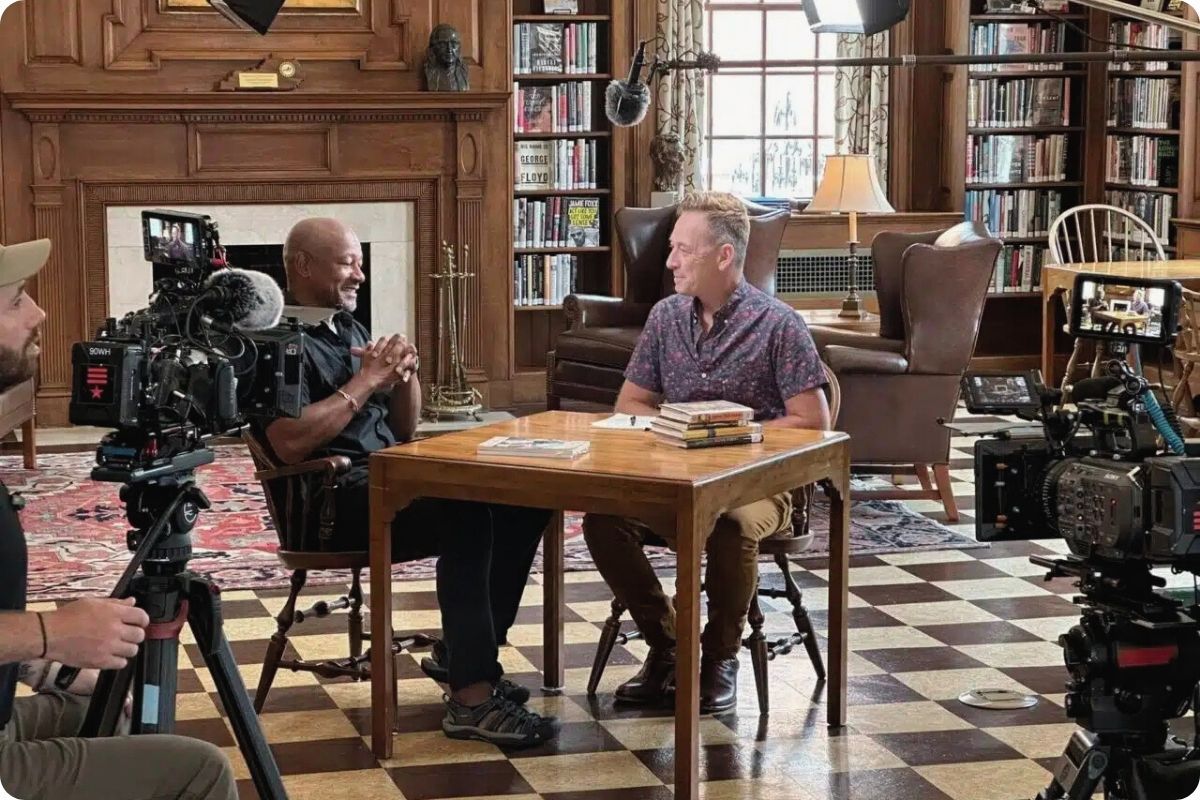 A behind-the-scenes look at a professional video interview. Two men sit at a wooden table in a classic library setting with floor-to-ceiling bookshelves, framed by cameras and production lighting.