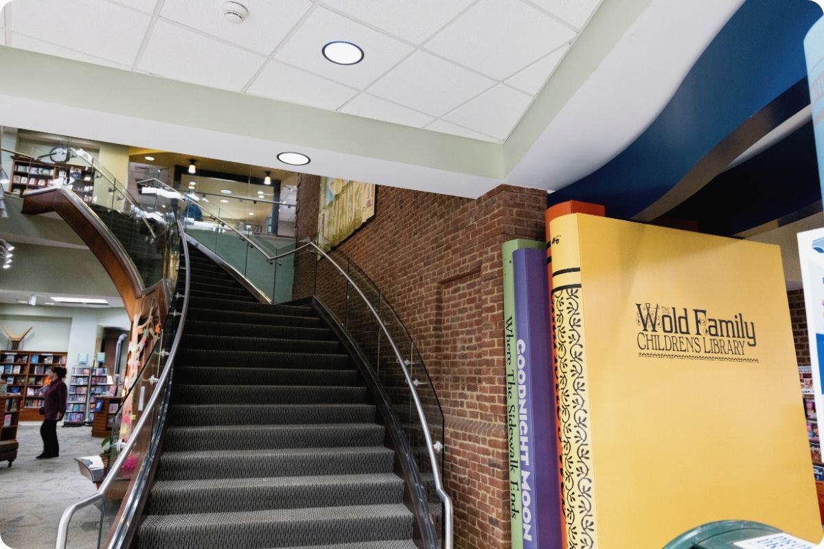 The interior of a modern library featuring a grand, curving staircase and a large yellow wall designed to look like a book spine for "The Wold Family Children’s Library."