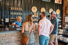 A bright, welcoming scene inside a distillery tasting room. A female staff member in a blue shirt smiles as she pours a sample for three customers at a rustic stone-faced bar. The background features barrels and shelves of bottled spirits.