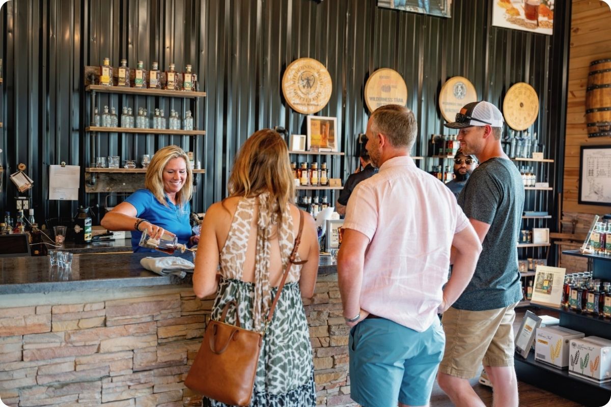 A bright, welcoming scene inside a distillery tasting room. A female staff member in a blue shirt smiles as she pours a sample for three customers at a rustic stone-faced bar. The background features barrels and shelves of bottled spirits.