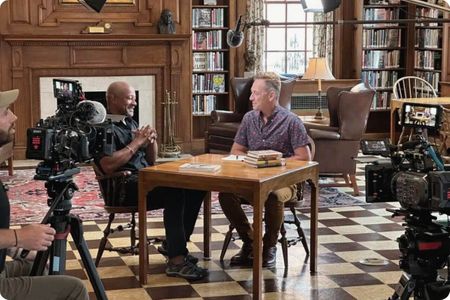 A behind-the-scenes view of a filmed interview in a classic library setting with two men seated at a table surrounded by professional cameras and lighting.