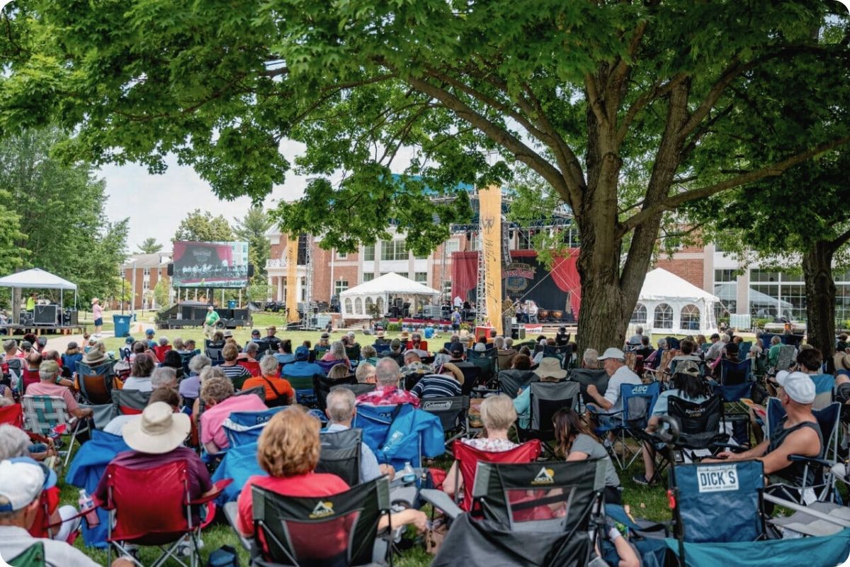 A wide shot of a large outdoor community event or festival. A diverse crowd of people sits in lawn chairs on a green campus, facing a large stage and video screen under the shade of mature trees.