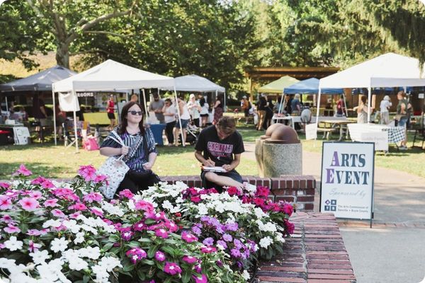 A lively outdoor community "Arts Event" held in a park. White pop-up tents are scattered across the grass, and people are seen browsing the booths, with a colorful flower bed of pink and white blossoms in the foreground.