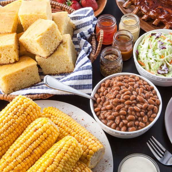 a table filled with cornbread, corn on the cob, baked beans, coleslaw and spices