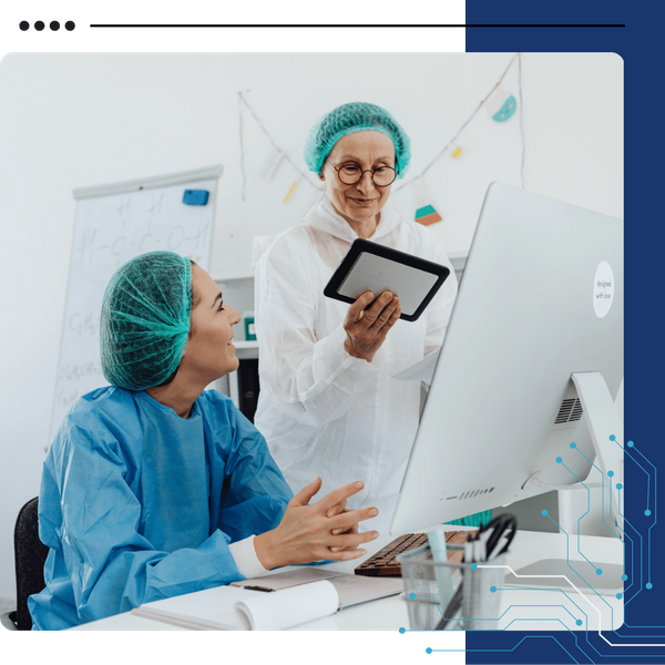 Two medical professionals in scrubs and hairnets looking at a computer screen and a tablet in an office setting.