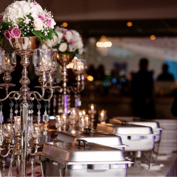 A close-up of a sophisticated buffet line for an event. Ornate silver candelabras with white and pink floral arrangements serve as centerpieces, alongside chafing dishes filled with hot food. Stacked plates are visible, and the background is softly blurred