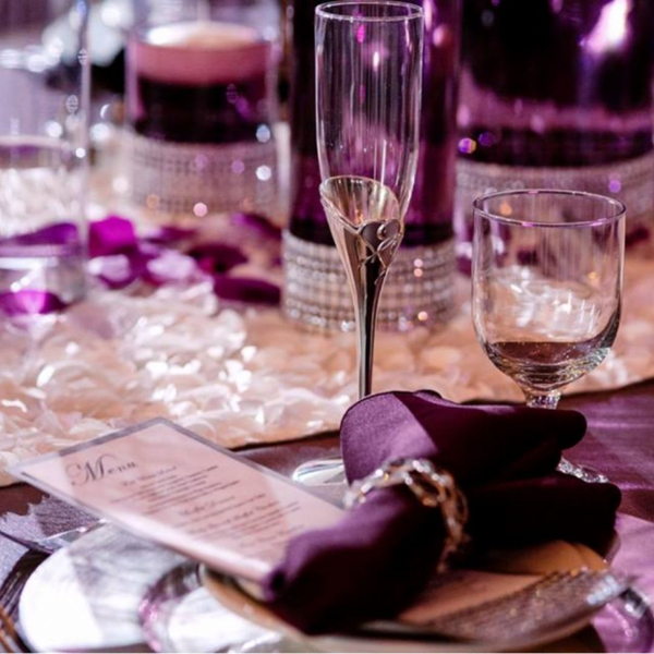 A close-up shot of an elegantly set table for an event, featuring purple and silver decor. A menu card is placed on a silver charger plate, next to a purple napkin with a decorative ring. A clear champagne flute and a water glass are visible, along with pu