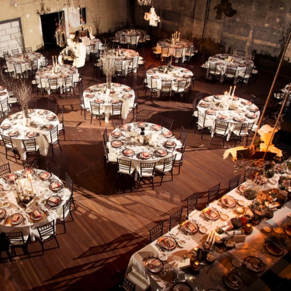 An overhead view of a beautifully set large event hall, likely a wedding reception, featuring numerous round tables with white tablecloths, chairs, place settings, and elegant centerpieces. The room has high ceilings and exposed industrial-style elements, 