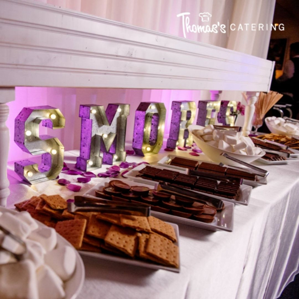 A festive dessert bar featuring S'mores ingredients. Large, illuminated marquee letters spell out "SMORES" in the background, with a purple glow. Plates of graham crackers, chocolate bars, and marshmallows are neatly arranged on a white tablecloth, ready f