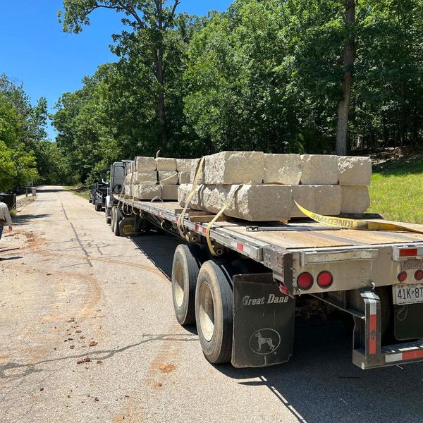 A flatbed semi-truck navigating a narrow road fully loaded with limestone blocks, highlighting our reliable wholesale rock logistics.