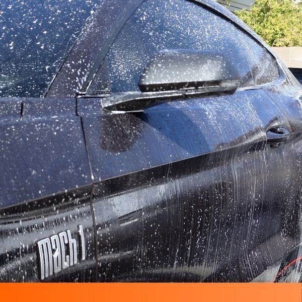 Side profile of a dark Ford Mustang Mach 1 covered in soap suds during a car wash, highlighting the "Mach 1" fender badge.