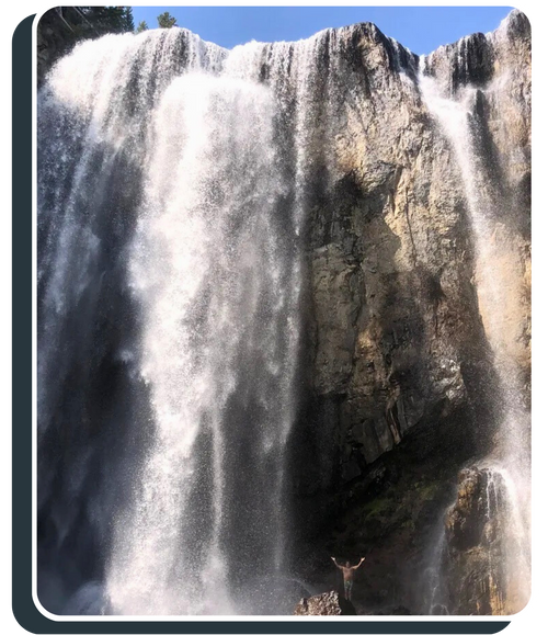 A large, powerful waterfall cascading over a rocky cliff. At the very bottom, a person stands on a rock with their arms raised in excitement, showing the massive scale of the falls.