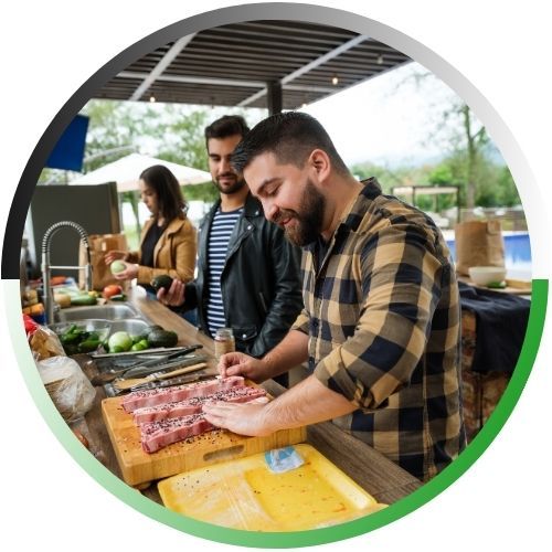 Circular icon showing friends gathered around a modern outdoor kitchen island while a man prepares seasoned meat for grilling.