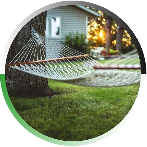A serene circular-framed view of a white rope hammock tied to a sturdy tree in a sun-drenched, well-maintained green lawn.