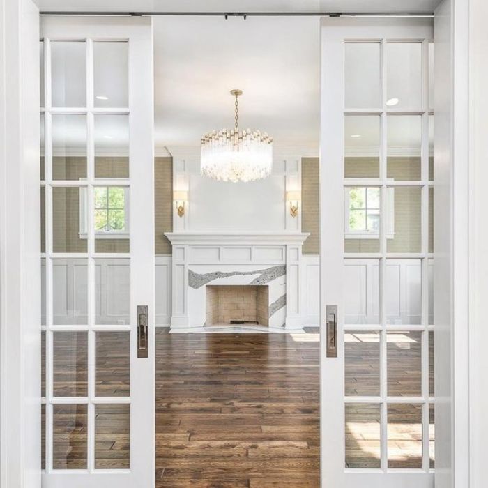 A view through open French doors into an elegant living room with a white fireplace. A tiered crystal chandelier hangs from the center of the ceiling. The room also features two small wall sconces with white candle-like lights on either side of the fireplace. A view through open French doors into an elegant living room with a white fireplace. A tiered crystal chandelier hangs from the center of the ceiling. The room also features two small wall sconces with white candle-like lights on either side of the firepla