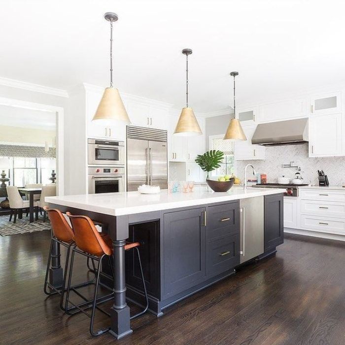 Three large, brushed-gold cone pendants hang over a dark gray kitchen island with leather stools in a spacious, white kitchen.