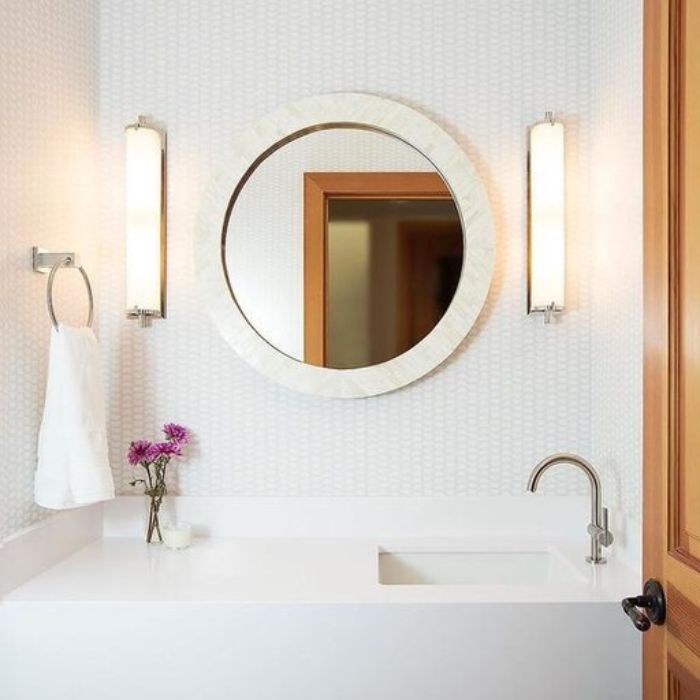 Two tall, cylindrical wall sconces with brushed nickel bases are mounted on either side of a large round mirror with a thick white frame. The sconces provide vertical lighting over a white bathroom vanity with a single-handle faucet. Two tall, cylindrical wall sconces with brushed nickel bases are mounted on either side of a large round mirror with a thick white frame. The sconces provide vertical lighting over a white bathroom vanity with a single-handle faucet.