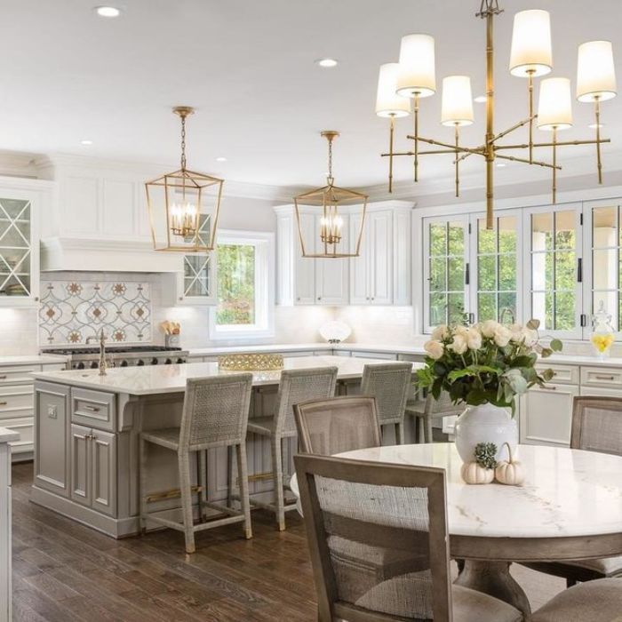 A modern kitchen with a large island and white cabinets. Two geometric brass pendants with exposed bulbs hang over the island. A multi-arm chandelier with white drum shades hangs over a round marble table in the foreground. A modern kitchen with a large island and white cabinets. Two geometric brass pendants with exposed bulbs hang over the island. A multi-arm chandelier with white drum shades hangs over a round marble table in the foreground.