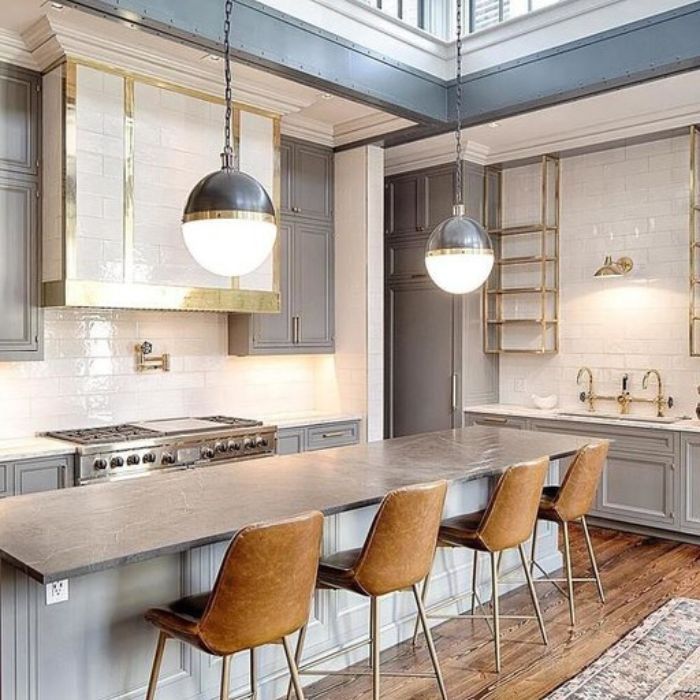 A modern kitchen with gray cabinets and gold hardware. Two large, spherical pendants with dark metal tops and frosted glass bottoms hang over a long kitchen island. A gold-toned decorative vent hood is centered above the range. A modern kitchen with gray cabinets and gold hardware. Two large, spherical pendants with dark metal tops and frosted glass bottoms hang over a long kitchen island. A gold-toned decorative vent hood is centered above the range.