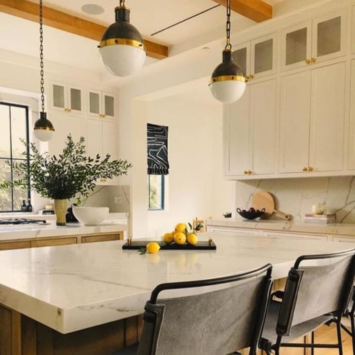 Large, brass-and-white globe pendants hang from chains over a marble-topped kitchen island in a bright kitchen with exposed wood beams. Large, brass-and-white globe pendants hang from chains over a marble-topped kitchen island in a bright kitchen with exposed wood beams.