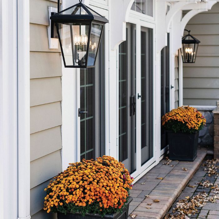 The exterior of a home with two black lantern-style outdoor sconces mounted on the wall. The sconces illuminate a brick walkway and a large planter filled with orange mums. The home features light-colored siding and white French doors. The exterior of a home with two black lantern-style outdoor sconces mounted on the wall. The sconces illuminate a brick walkway and a large planter filled with orange mums. The home features light-colored siding and white French doors.