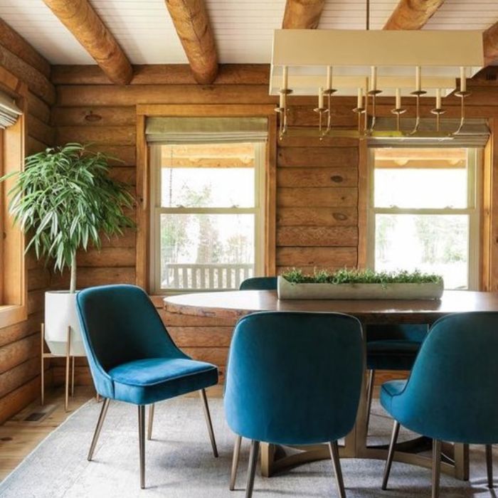 A modern rectangular chandelier hangs above a dining table with teal chairs in a rustic log cabin dining room. A modern rectangular chandelier hangs above a dining table with teal chairs in a rustic log cabin dining room.