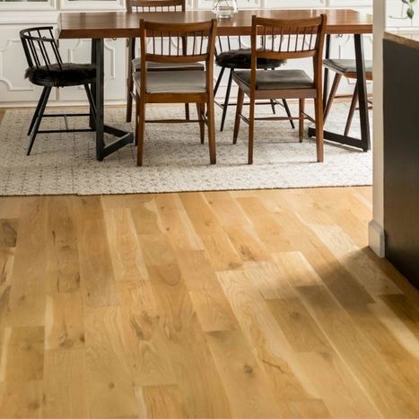 Wooden dining room table and chairs on a textured white area rug over light wood-look flooring.