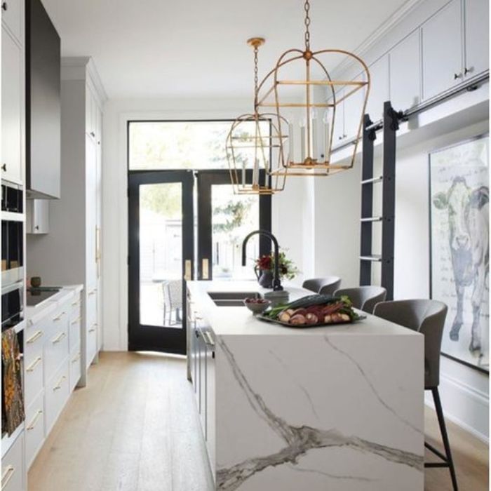 Two delicate, gold geometric cage pendants hang over a marble waterfall island in a narrow, modern galley kitchen. Two delicate, gold geometric cage pendants hang over a marble waterfall island in a narrow, modern galley kitchen.