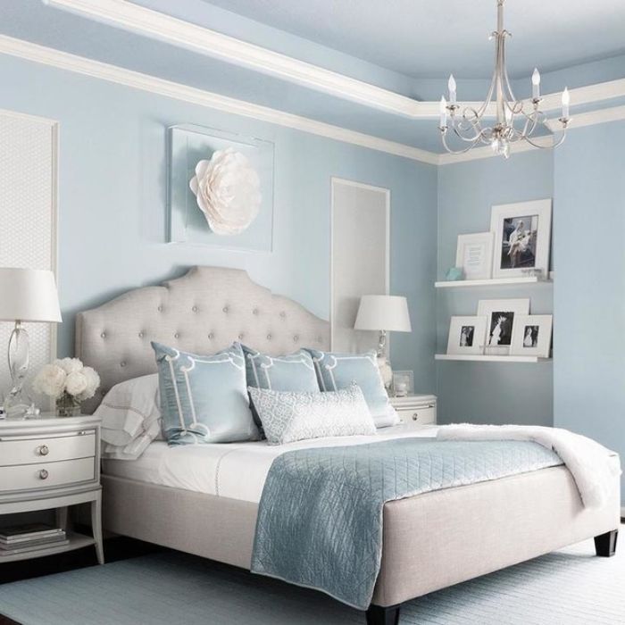A large, elegant crystal chandelier hangs from a blue ceiling in a serene bedroom. A white tufted headboard is centered under a decorative ceiling medallion. Two table lamps with white shades sit on matching bedside tables. A large, elegant crystal chandelier hangs from a blue ceiling in a serene bedroom. A white tufted headboard is centered under a decorative ceiling medallion. Two table lamps with white shades sit on matching bedside tables.
