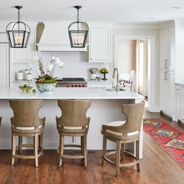 Two large, black, lantern-style pendants hang over a white kitchen island featuring three woven-back bar stools. Two large, black, lantern-style pendants hang over a white kitchen island featuring three woven-back bar stools.